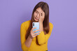 © sementsova321 - Face palm from stupid news, upset young woman with long hair, covering face with hand, holding smart phone, standing isolated over lilac background, dresses casual attire.