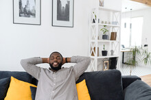 Satisfied young african man resting on comfortable couch in white cozy modern living room, holding hands behind head, millennial hipster guy enjoy no stress life on sofa at home with his eyes closed
