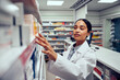 © StratfordProductions - Young woman working in pharmacy looking for medicine in shelf standing behind counter