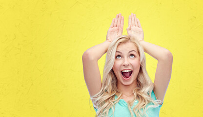 Wall Mural - fun, expressions, easter and people concept - happy smiling young woman making bunny ears over yellow background