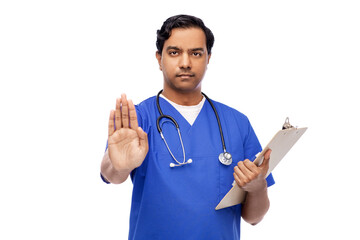 Wall Mural - healthcare, profession and medicine concept - indian male doctor in blue uniform with stethoscope and clipboard showing stop gesture over white background