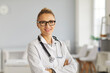 © Studio Romantic - Portrait of friendly doctor or nurse at work. Professional medical staff headshot. Happy young woman in eyeglasses and white lab coat with stethoscope standing arms crossed and smiling at camera