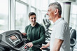 Close up in focus of an older man's face, he is looking ahead and performing a cardio exercise, in the background his rehabilitation coach