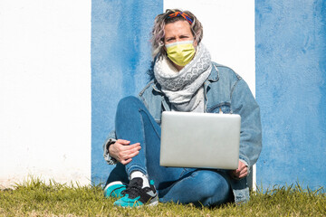 Poster - Young Caucasian female with a face mask sitting against a blue and white wall working on the laptop