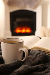 Sticker - Cup of hot tea and book on knitted plaid near fireplace at home. Cozy atmosphere