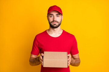 Wall Mural - Photo portrait of serious confident courier keeping carton box for delivery in red uniform cap isolated on bright yellow color background