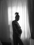Fototapeta Młodzieżowe - The silhouette of a pregnant girl standing by the window. Black and white photo.