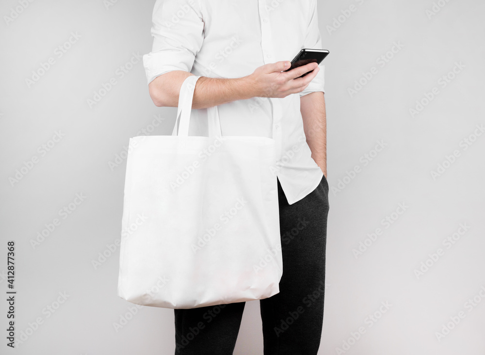 a man stands on a white background, reads the news on the phone and holds an ecological bag made of flax on his elbow. Ecology Concept - obrazy, fototapety, plakaty 