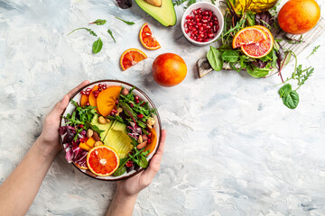 Wall Mural - Girls' hands holding vegan, detox Buddha bowl with avocado, persimmon, blood orange, nuts, spinach, arugula and pomegranate on a light background, top view
