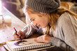 © Mint Images - Teenage girl in a woolly hat drawing with a pencil on a sketchpad.
