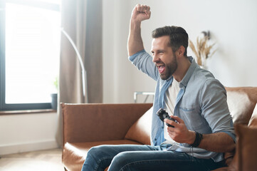 Side view a gambling guy with a joystick sitting on the couch, scream happily, won soccer on console. A bearded man spends leisure with a video game