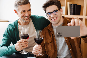 Wall Mural - Happy gay couple drinking wine while taking selfie on cellphone