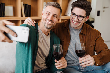 Wall Mural - Happy gay couple drinking wine while taking selfie on cellphone