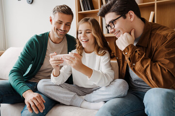 Wall Mural - Happy gay family smiling and using mobile phone while sitting on couch