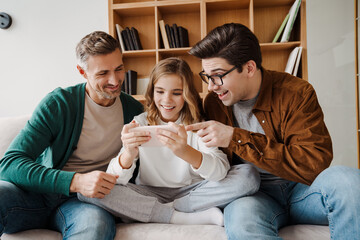 Wall Mural - Happy gay family smiling and using mobile phone while sitting on couch