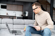 © Vadim Pastuh - A male employee is working with laptop remotely from home, a guy is looking through workpapers, analyzing tasks, researching startup ideas. Freelancer works on the distance
