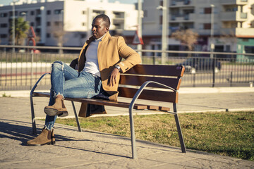 Wall Mural - Closeup shot of a stylish handsome African male in a coat sitting on a bench