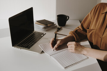 Wall Mural - Minimalist home office workspace with laptop, coffee cup, clipboard. Woman write in a paper sheet notebook. Morning work, business concept. Sunlight shadows on the wall.