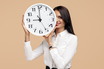 Photo of young modern business woman hold a clock in front of the face over beige background.