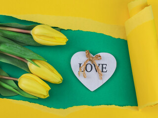 Wall Mural - card with tulips and heart.
Three tulips and a wooden heart in a yellow paper torn frame on a green background, top view close-up.