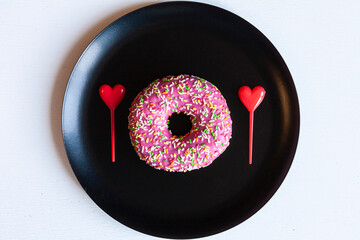 a pink doughnut and two red plastic hearts on a black plate on white background