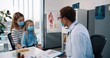 © VAKSMANV - Portrait of African American young happy male professional physician in white medical coat speaking on consultation with pretty Caucasian mother and little daughter girl in medical mask, healthcare