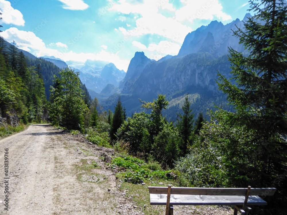 Gravelled road in the mountains with the view on high Alps in the region of Gosau, Austria. A wooden bench next to the road, with the wie on Gosau lake. Endless mountain chains. Serenity and calmness - obrazy, fototapety, plakaty 