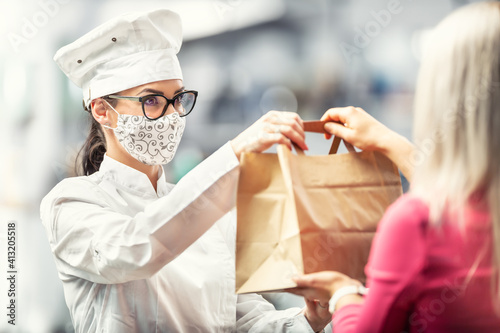 Chef wearing hat, glasses and face mask hands a paper bag with delivery food to a customer © weyo
