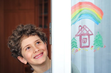 Cute little boy with a rainbow and a house painted on the window during quarantine due to the coronavirus pandemic.