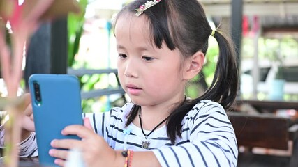 Poster - Asian Little girl hand holding and watching playing game in smartphone at cafe. kid addicted to games problems of children use smartphone technology Natural light outdoor lifestyle