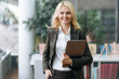 © Kateryna - Formal portrait of successful elegant business lady dressed in suit, stands in modern office, holding laptop in arm. Charming mature blonde woman with looking and smiling directly at the camera