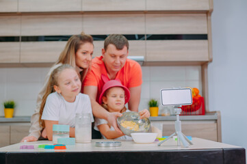 Wall Mural - Mom with Father teaches two daughters to cook dough in the kitchen. The family is filming a culinary video on a smartphone.