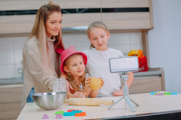 Wall Mural - Mom teaches her daughters to cook dough in the kitchen. The family is filming a culinary video on a smartphone.