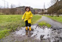 Portrait Of A Little Boy Smiling And Jumping In A Puddle Of Rainwater In A Muddy Puddle On A Meadow Road Wearing A Yellow Raincoat And Yellow Rain Boots