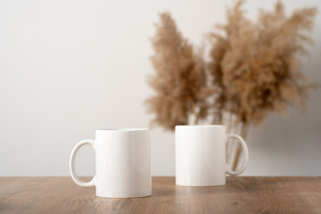 Mockup two white mugs on a wooden table top with pampas decor in Scandinavian interior, boho style. Template, layout for your design, advertising, logo with copy space. Cups on light beige background