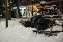 Homeless Man Sleeping On Snowy Bench In Park