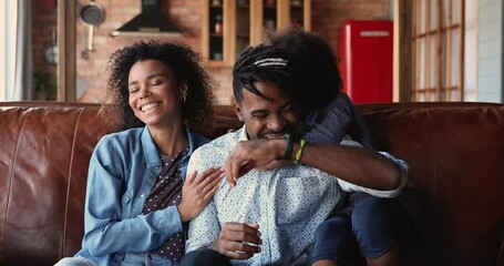 Wall Mural - Happy small african ethnicity child girl tickling, playing with joyful loving biracial parents, sitting together on comfortable couch, overjoyed mixed race family having fun together in living room.