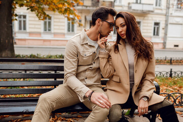 Wall Mural - Brunette model with stylish guy in beige coat sitting on bench.