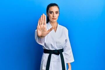 Wall Mural - Beautiful brunette young woman wearing karate fighter uniform with black belt doing stop sing with palm of the hand. warning expression with negative and serious gesture on the face.