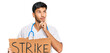 © Krakenimages.com - Young handsome man wearing doctor uniform holding strike banner cardboard serious face thinking about question with hand on chin, thoughtful about confusing idea