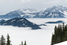 The Valley Of Winter Kitzbuhel Is Covered With Clouds. Mountain Peaks And A Coniferous Forest Rise Out Of The Fog.