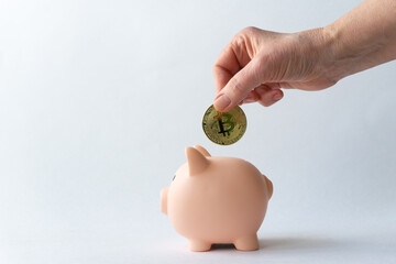 Hand of elderly Caucasian woman drops metal bitcoin coin into piggy bank pink pig on white background with place to place text