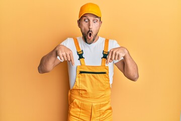 Young handsome man wearing handyman uniform over yellow background pointing down with fingers showing advertisement, surprised face and open mouth