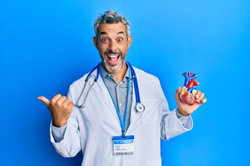 Middle age grey-haired man wearing doctor uniform holding heart pointing thumb up to the side smiling happy with open mouth