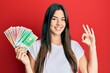 © Krakenimages.com - Young brunette woman holding norwegian krone banknotes doing ok sign with fingers, smiling friendly gesturing excellent symbol