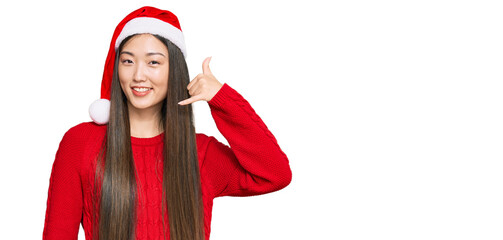 Young chinese woman wearing christmas hat smiling doing phone gesture with hand and fingers like talking on the telephone. communicating concepts.