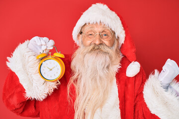 Wall Mural - Old senior man with grey hair and long beard wearing santa claus costume holding alarm clock screaming proud, celebrating victory and success very excited with raised arm