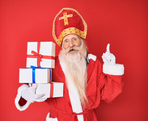 Wall Mural - Old senior man with grey hair and long beard wearing saint nicholas costume holding presents surprised with an idea or question pointing finger with happy face, number one