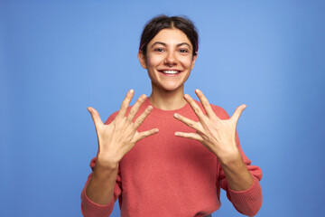 Wall Mural - Cute happy teenage girl with nose ring looking at camera with excited radiant smile, holding hands, spreading fingers wide, showing neat manicured nails, posing isolated against blue background