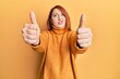 Beautiful redhead woman doing thumbs up positive gesture in shock face, looking skeptical and sarcastic, surprised with open mouth